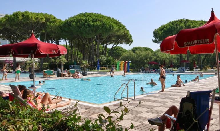 Outdoor pool with red umbrellas and people relaxing.
